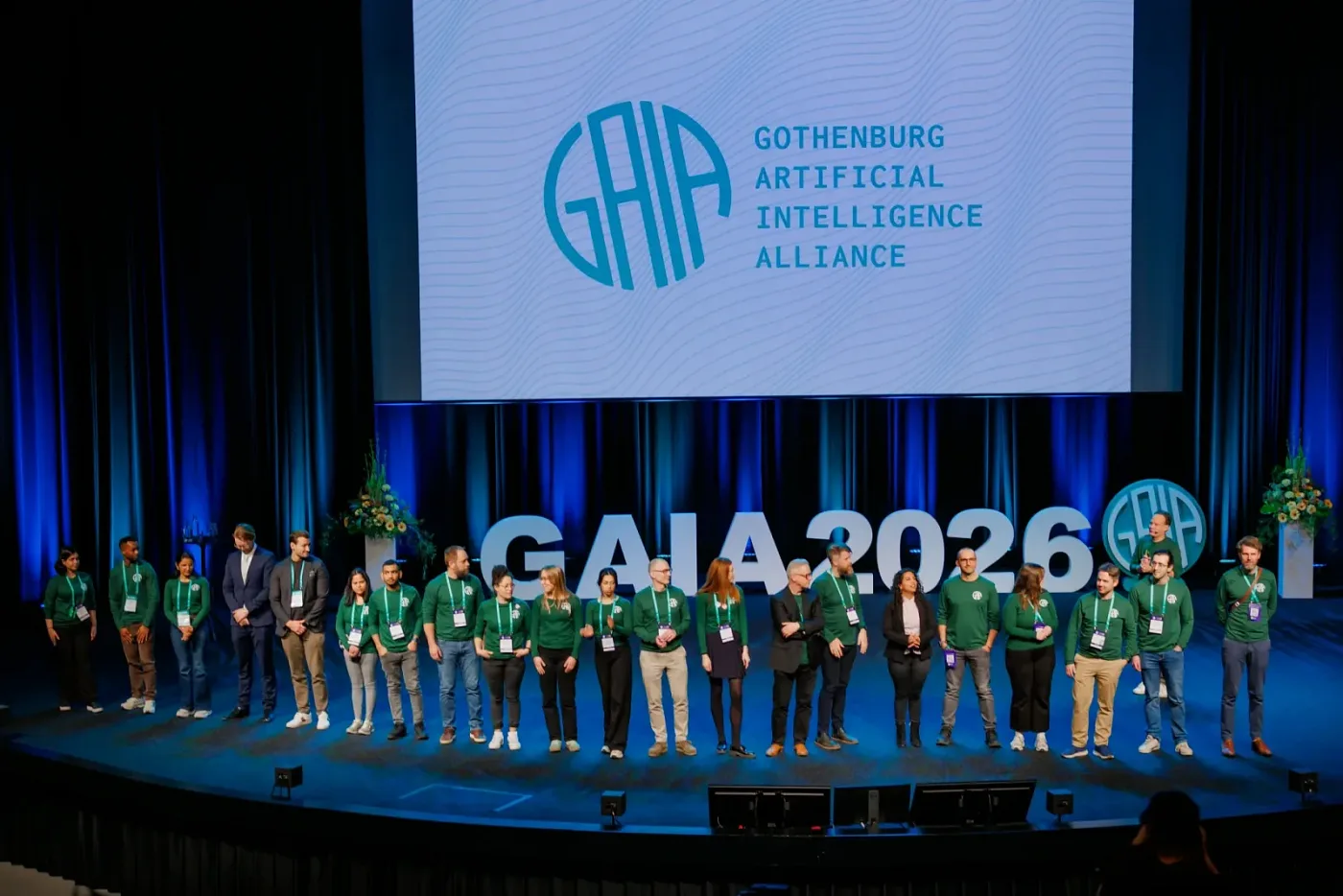 Organising team in green sweaters on stage in front of the GAIA 2026 logo
