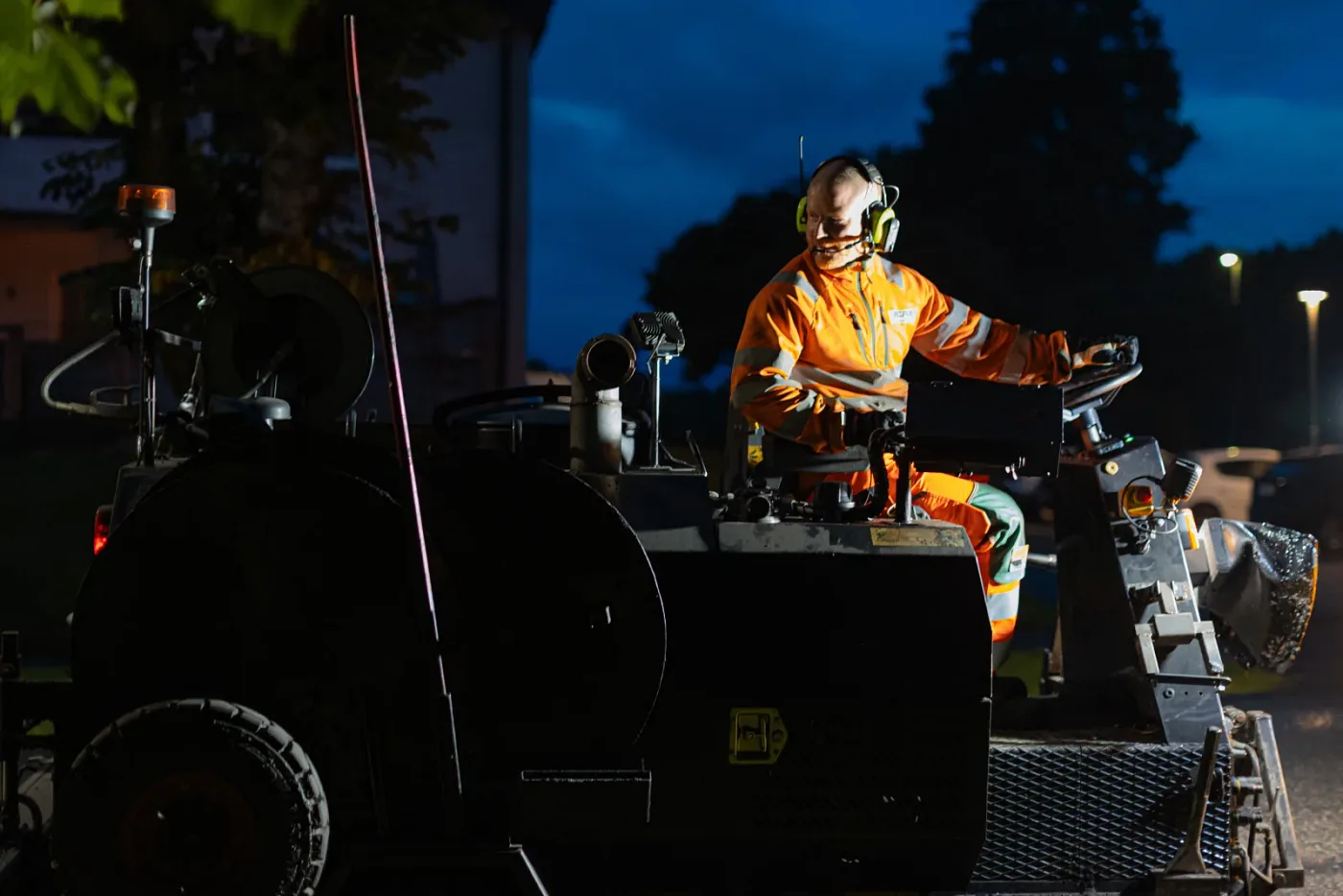 Machine operator in hi-vis clothing and ear defenders at the controls of an asphalt machine in the evening