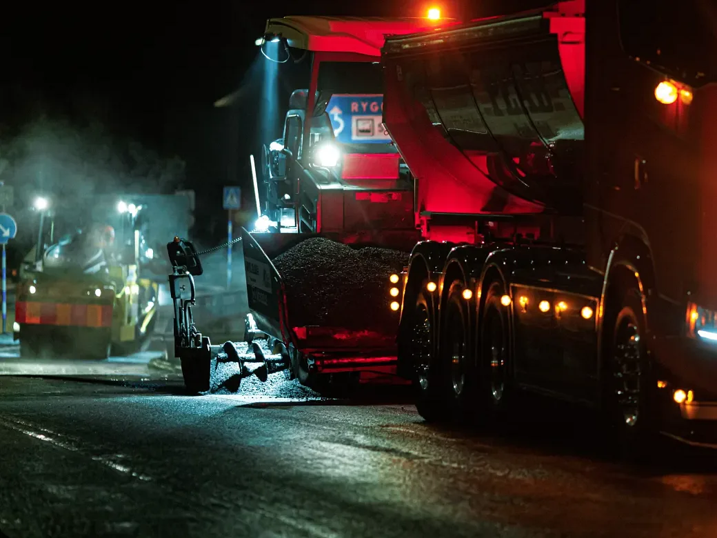 Asphalt paver being loaded with hot asphalt from a truck under floodlights at night