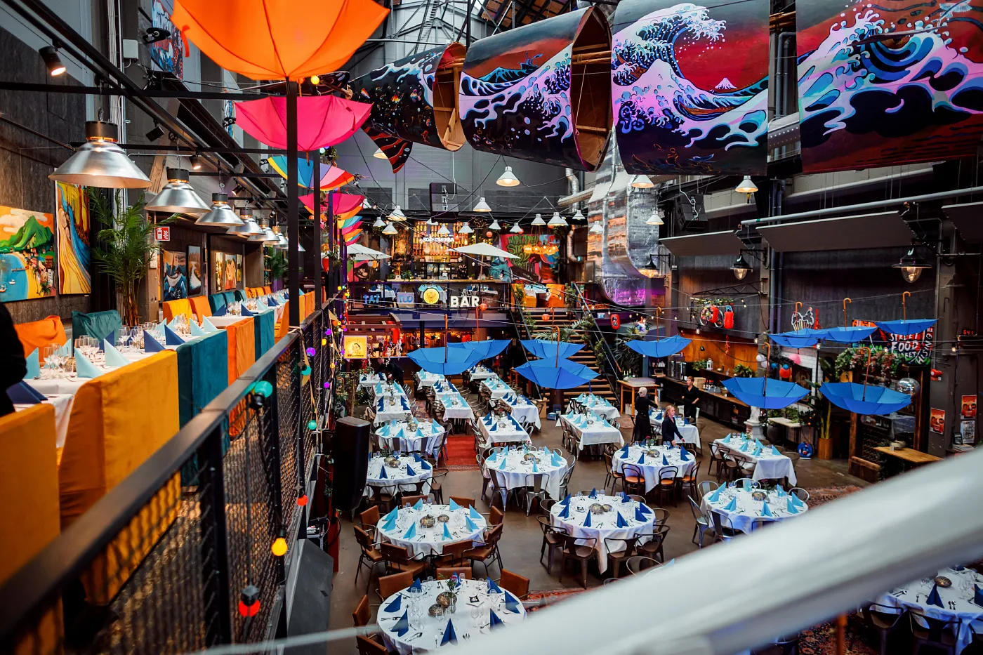 A large hall with laid tables under blue parasols, seen from the balcony