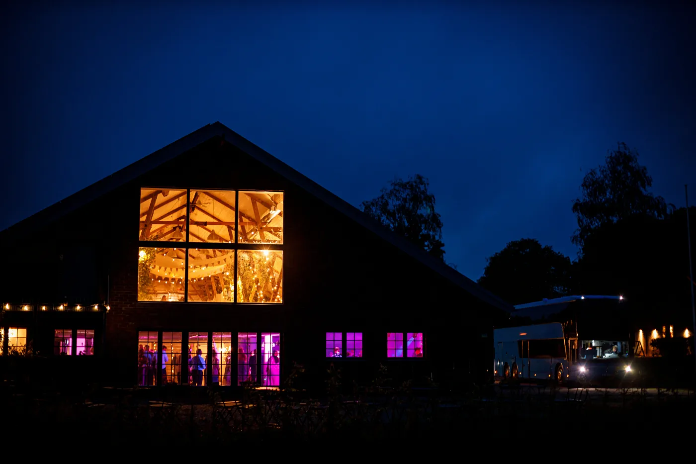A countryside venue at dusk: warm light in the windows and guest silhouettes inside