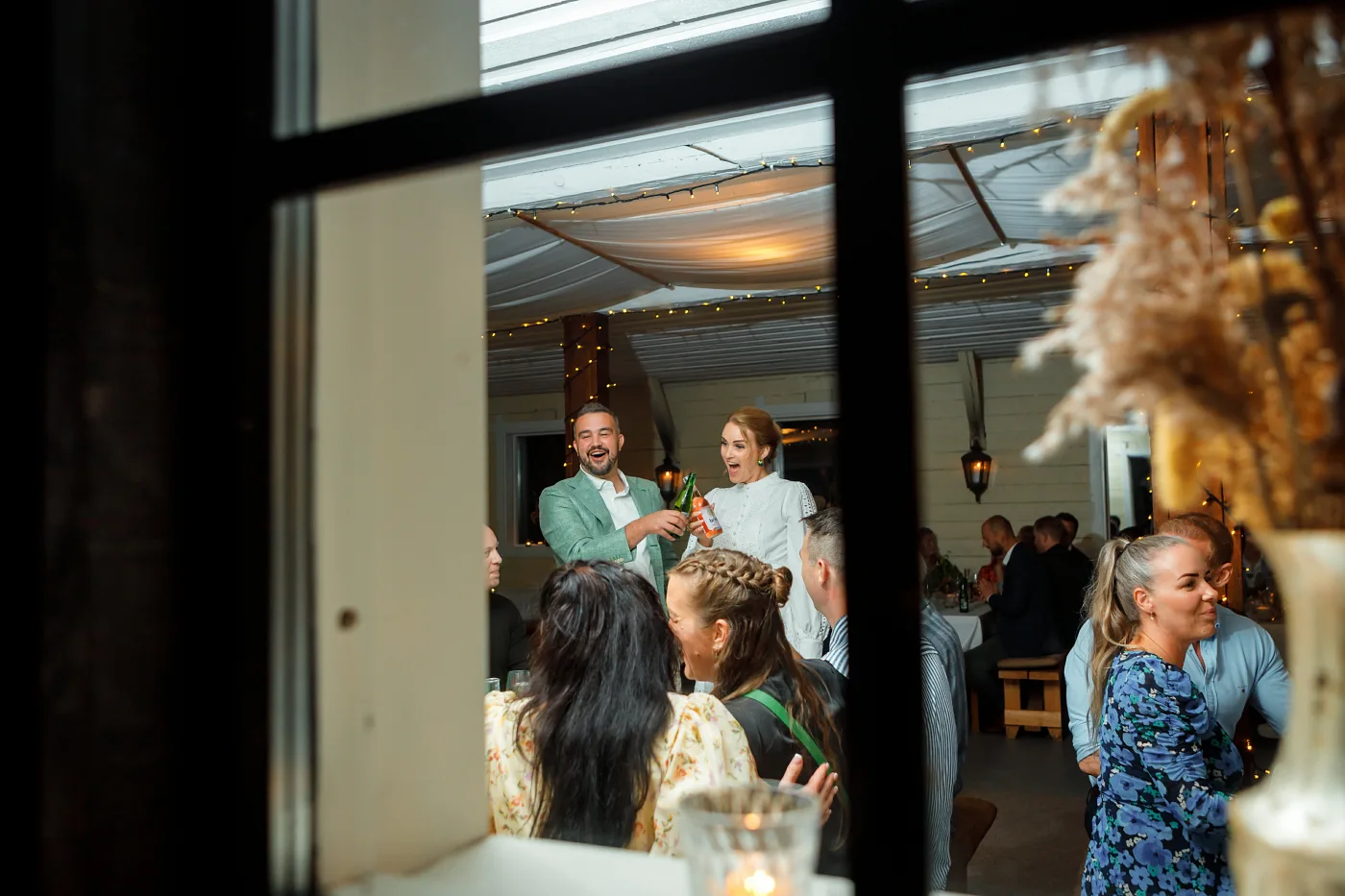 Shot through a window: the couple standing among the guests under string lights, everyone laughing