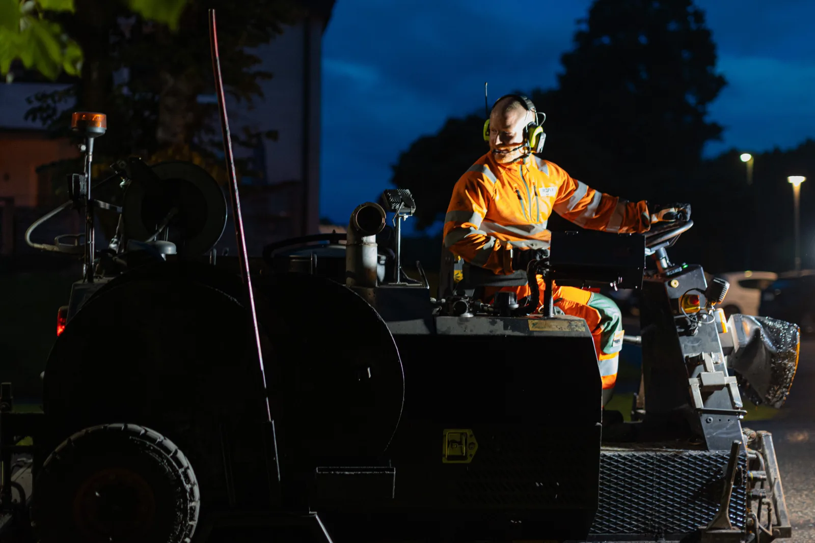 Machine operator in orange high-visibility clothing and ear defenders at blue dusk