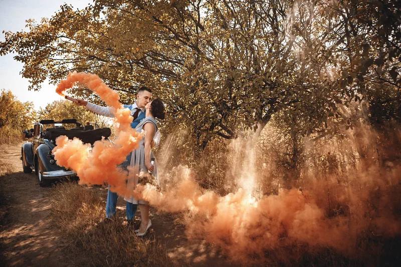 Bride and groom kissing beside a vintage car in orange smoke